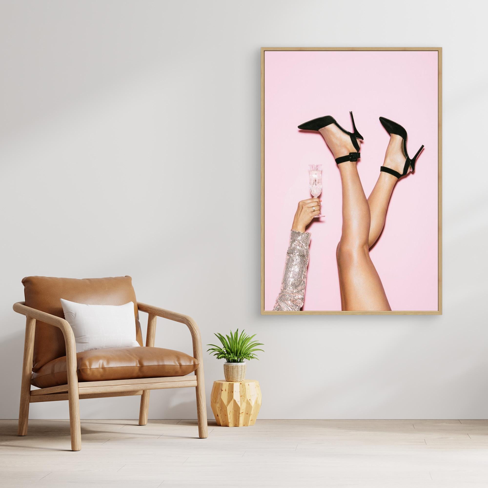 Framed artwork of legs in high heels holding a glass on a pink background, placed above a brown armchair.