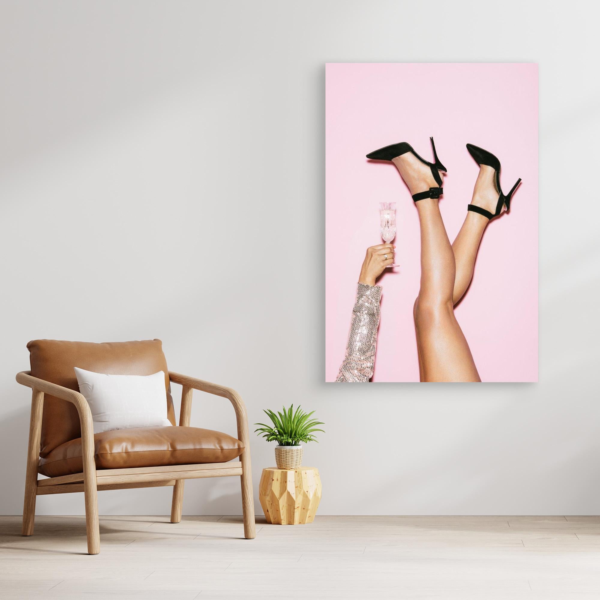 Canvas print of legs in high heels holding a glass against a pink background, placed on a wall above a brown armchair.